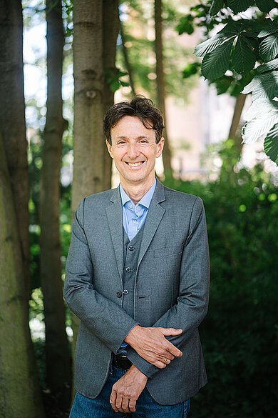 Prof. Dr. Michael Jaensch stands in the shade of trees and bushes wearing a grey jacket and light blue shirt 
