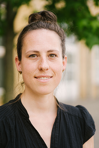 Porträt von Elise Schwarz. Eine lächelnde Frau. Sie trägt eine schwarze Bluse und hat die dunklen Haare zu einem hohen Dutt gebunden.