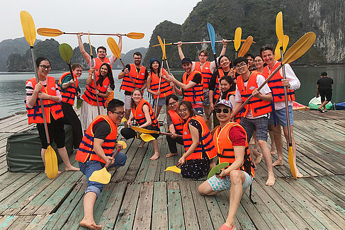 Reisegruppe steht in orangefarbenen Schwimmwesten und mit Paddeln in den Händen für ein Gruppenfoto auf einem Steg vor der Kulisse der Halong-Bucht
