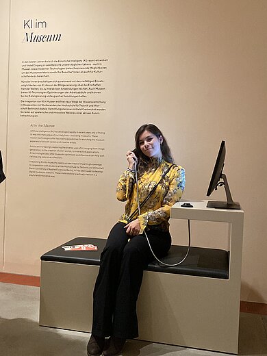 Sophie sitzt im Staatlichen Museum Schwerin an einer interaktiven Station der Ausstellung ‚KI im Museum‘ und führt ein Gespräch über ein Telefon; neben ihr befindet sich ein Bildschirm.