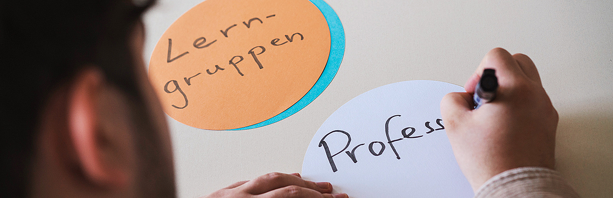 Ein Studierender sitzt am Tisch und beschriftet bunte Pappkarten. Auf einer Karte steht "Lerngruppen". Die zweite wird gerade beschriftet und es ist "Profess" zu lesen.