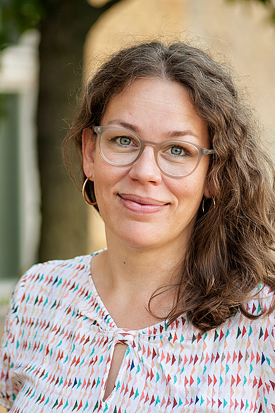 Porträt von Nadja Bajerski. Eine lächelnde Frau mit dunkelblonden, schulterlangen Locken und einem hellen, gemusterten Shirt. Sie trägt eine Brille.