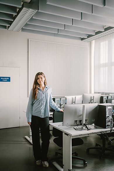 Sophie steht in einem Computerraum der HTW Berlin neben einem Arbeitsplatz mit mehreren Monitoren; der Raum ist hell und modern eingerichtet.