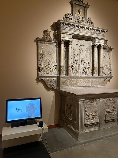 Steinernes, reich verziertes Altarrelief mit Säulen und biblischen Szenen neben einem Monitor auf einem weißen Tisch in einem Museum.