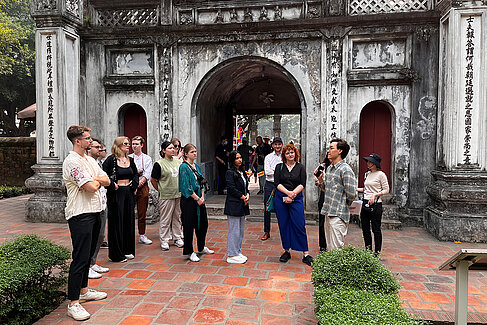 The tour group is standing in front of a large grey stone gate decorated with Asian lettering in the grounds of the Temple of Literature and is listening to someone speak