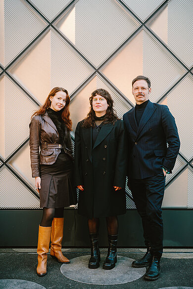 Drei Personen des Projektteams stehen vor einer Wand mit geometrischem Muster, zwei Frauen tragen lange Mäntel und Stiefel, ein Mann trägt einen dunklen Anzug und schwarze Schuhe.