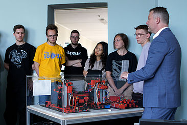 Professor Wittenberg und Studierende stehen um die Minifabrik herum. Der Professor zeigt und erklärt den Studierenden die Funktionsweise der Anlage.
