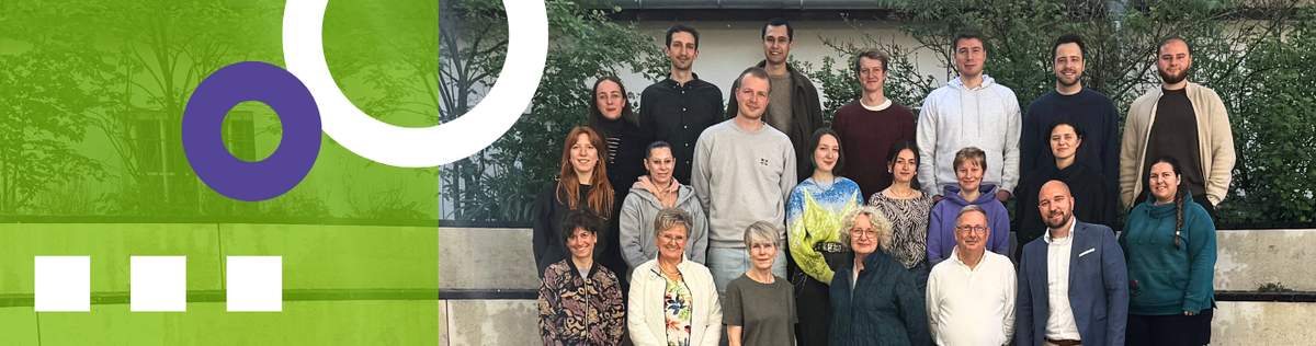 Gute Zusammenarbeit © Charlotte Meusinger Eine Gruppe von Menschen steht in zwei Reihen vor einer niedrigen Betonmauer im Freien. Im Hintergrund sind grüne Sträucher und ein Gebäude mit großen Fenstern zu sehen. Links im Bild befindet sich ein halbtransparenter, grüner Bereich mit weißen und lilafarbenen grafischen Elementen.