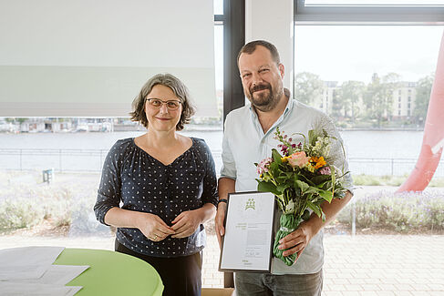 Zwei Personen stehen nebeneinander in einem hellen Raum mit Blick auf einen Fluss. Der Mann, Stephen Mortimer, hält einen Blumenstrauß und eine gerahmte Urkunde mit der Aufschrift „Preis für gute Lehre 2025“. Die Präsidentin der HTW, Annabella Rau