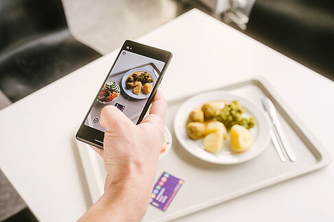 Ein Hand mit einem Handy, daneben ein Mensatablett mit Essen