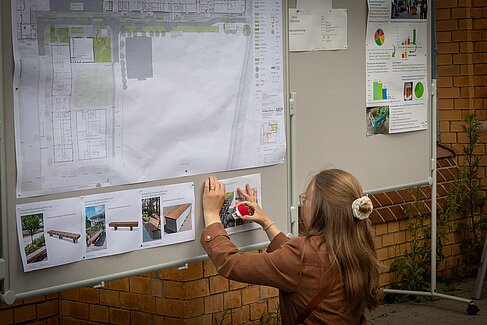 Pinnwand mit Plönen, davor eine junge Frau, die etwas an der Pinnwand befestigt