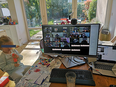 Bildschirm und Tastatur auf einem Tisch, an dem auch ein Mädchen mit Malsachen sitzt