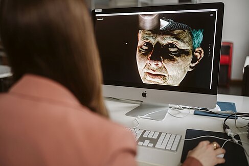 Chantal Pisarzowski am Computer, auf dem Monitor das virtuelle Gesicht einer alten Frau