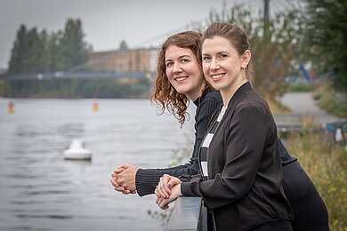 Sarah Marx und Anne Hübinger lehnen an einem Geländer am Flussufer und lächeln in die Kamera.