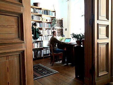 Eine Tür gibt den Blick in einen Raum frei, in dem ein Mann am Schreibtisch sitzt.