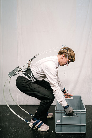 Mann mit Exoskelett bückt sich und hält eine Kiste mit beiden Händen